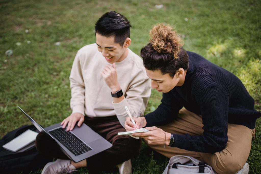 From above happy ethnic male students in casual wear using laptop and taking notes in copybook while preparing for exams together and sitting on grass in lush park